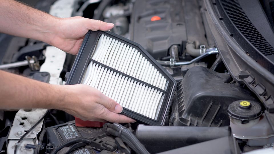 a man holding a air filter in front of a car.|a man in a white shirt and blue gloves working on a car engine.