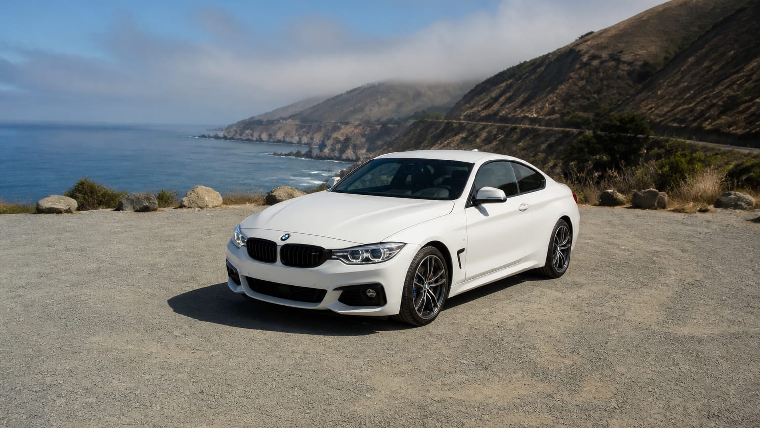 A white BMW 4 Series coupe parked on a coastal canyon overlook.