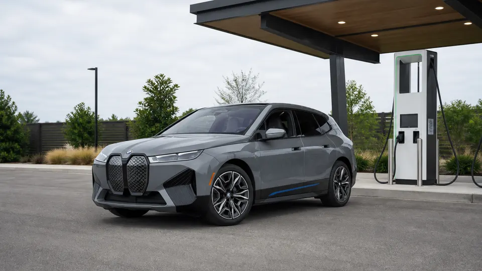 A Sophisto Grey BMW iX electric SUV, three-quarter front view, parked beside a modern charging plaza.