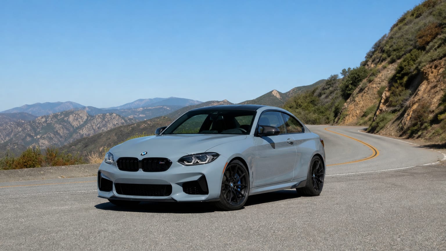 A late-model 2020–2021 or 2024 BMW M2 on a canyon road.