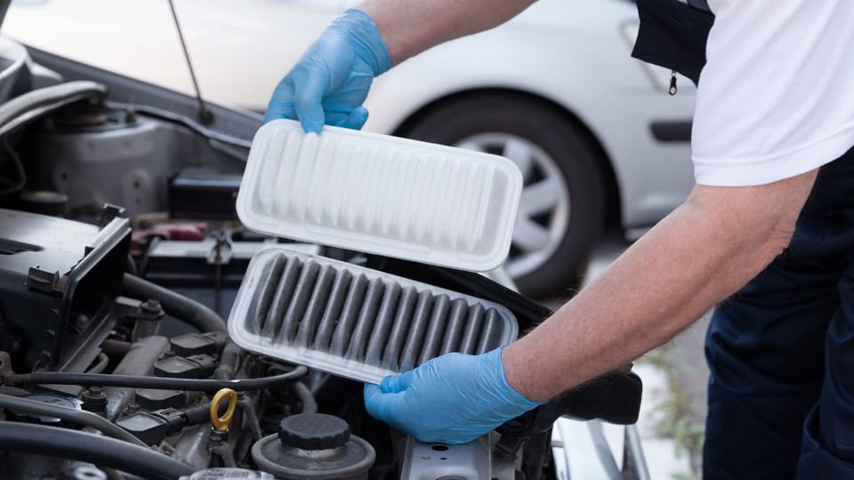 a man in a white shirt and blue gloves working on a car engine.|the engine of a red car with a filter on it.|a mechanic working on a car engine.