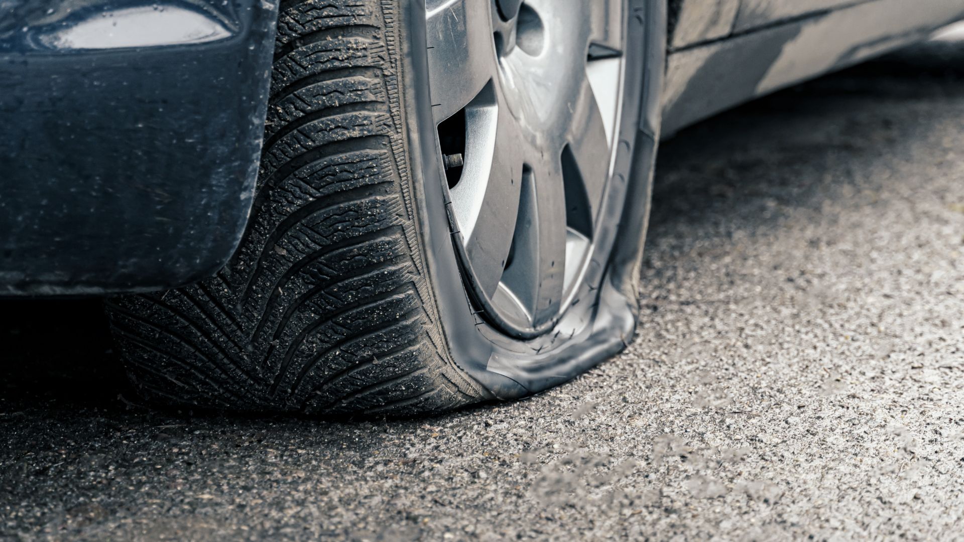 a man kneeling down next to a car holding a cell phone.|a close up of a car tire on a wet road.|a close up of a speedometer on a car.|a man fixing a tire on the side of a road.