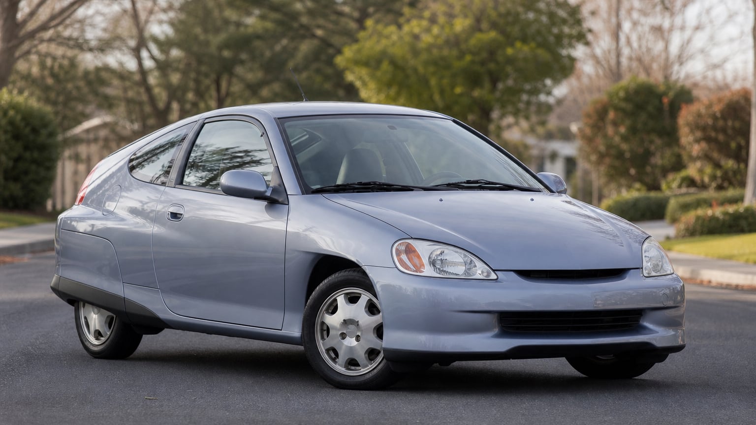 A silver-blue first-generation Honda Insight hybrid coupe parked on a suburban street, showing its teardrop body and rear wheel skirts.