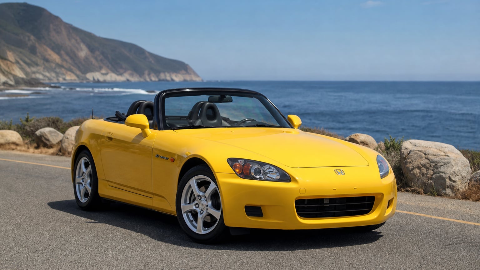 A yellow Honda S2000 roadster with the soft top down parked on a coastal road.