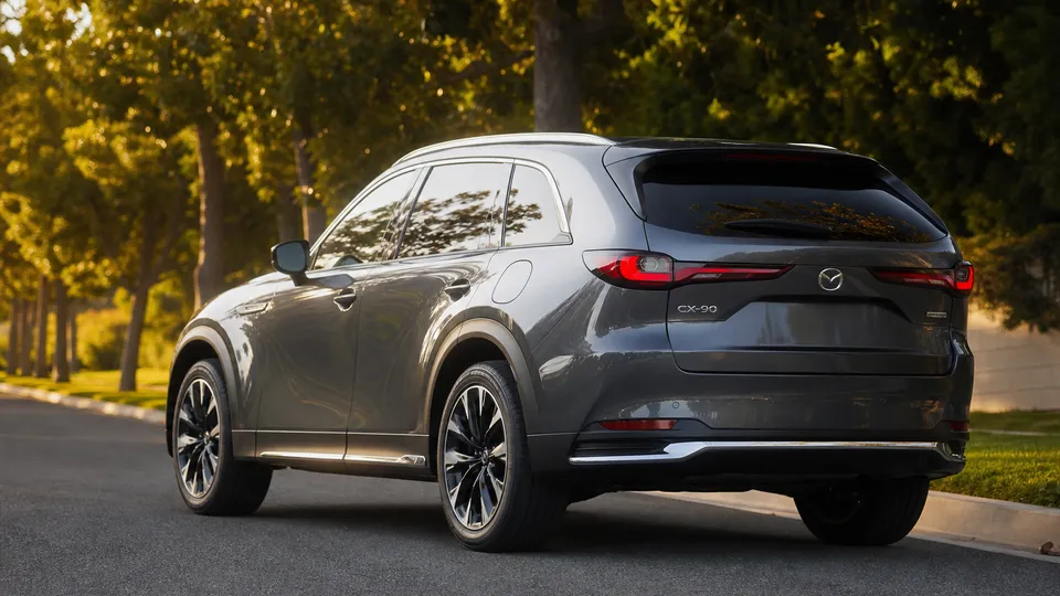 A gray Mazda CX-90 three-row SUV, rear three-quarter view, on a tree-lined street.